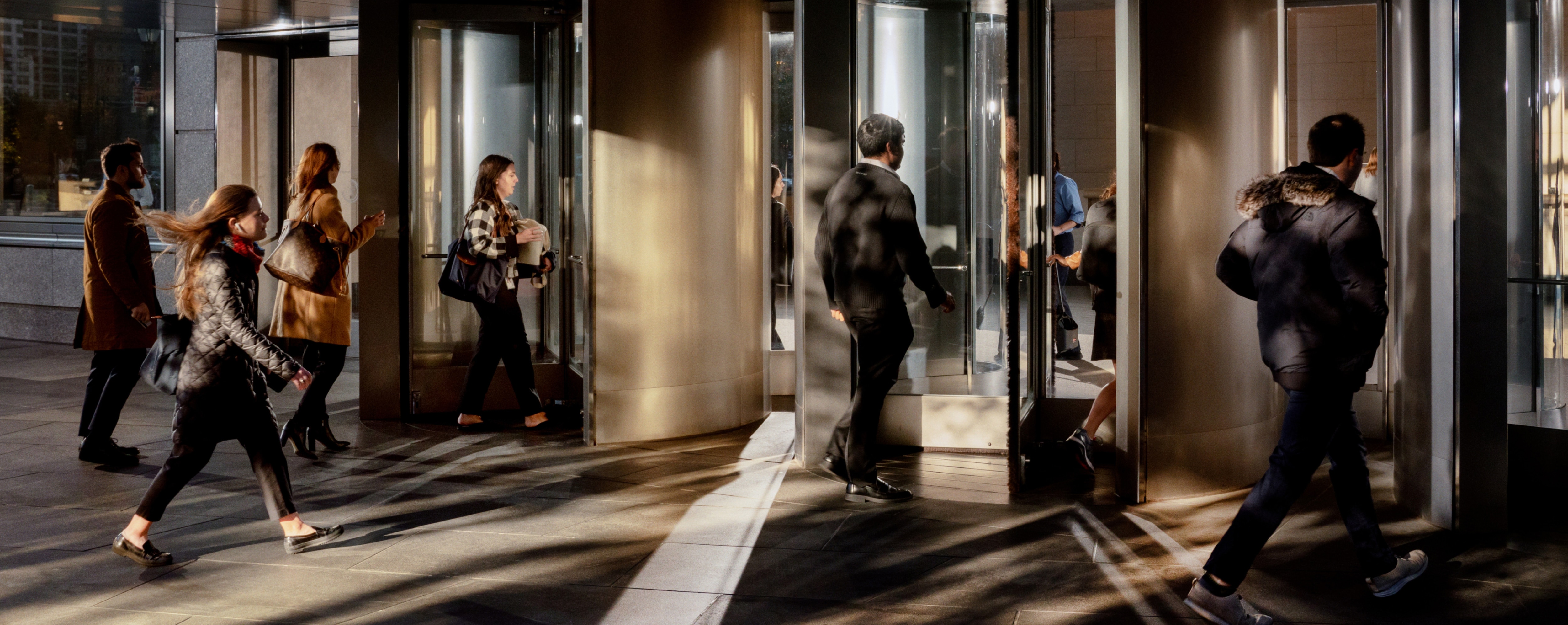 People entering and exiting a building through revolving doors in the early morning light.