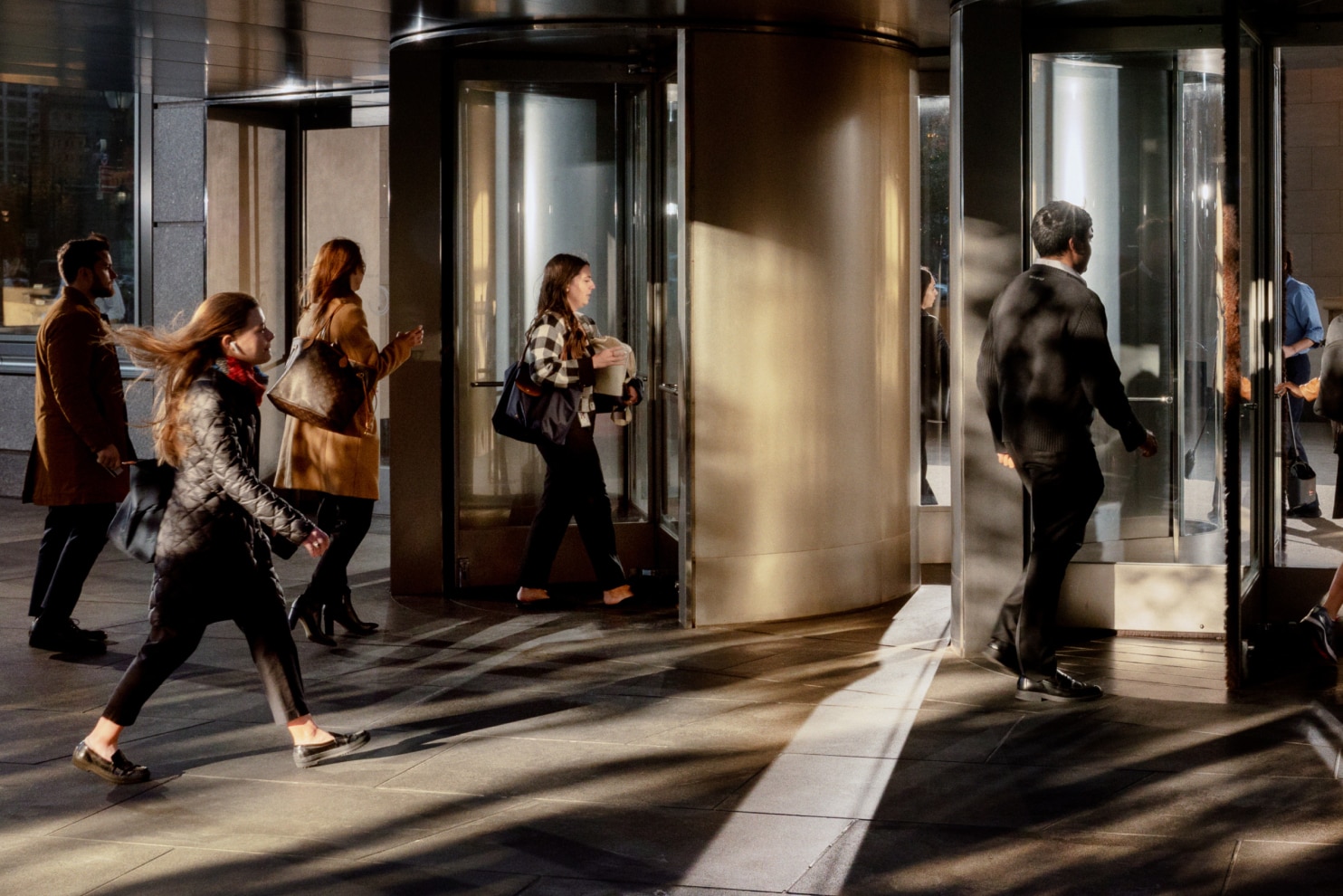 Three professionally dressed individuals walk through a stylish, modern lobby with curved architectural elements. The man on the left, wearing a green sweater and dark pants, carries a brown leather duffle bag. The woman in the middle wears a striped blouse with a sweater draped over her shoulders and carries a black handbag. The woman on the right, dressed in a navy blue top and wide-leg trousers, pulls a sleek silver rolling suitcase while holding a phone. They appear engaged in a friendly conversation in a travel or business setting.