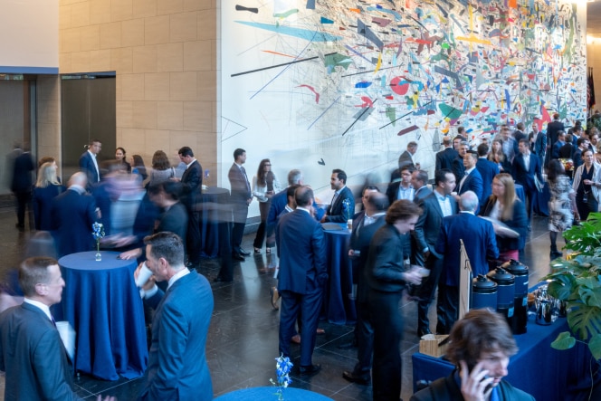 A large group of professionals networking in a spacious venue with high ceilings, blue tablecloth-covered cocktail tables, and a vibrant abstract mural on the wall.