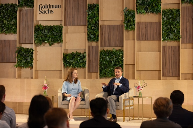 A woman and a man warmly shaking hands at a networking event, both smiling, with blurred figures in the foreground suggesting a busy environment.