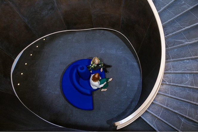 A high-angle shot captures a spiral staircase with two women seated on a blue, snail-shaped couch. The staircase features dark gray walls and gray steps with a white handrail. Small lights line the wall along the spiral. The women, one with blonde hair and one with red hair, are facing each other.