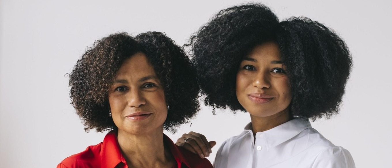 Edith Cooper and Jordan Taylor standing side by side against a plain white background. Edith, on the left, wears a red blouse and smiles softly, while Jordan, on the right, wears a white shirt and smiles warmly. Both have natural curly hair and exude a sense of connection and confidence.