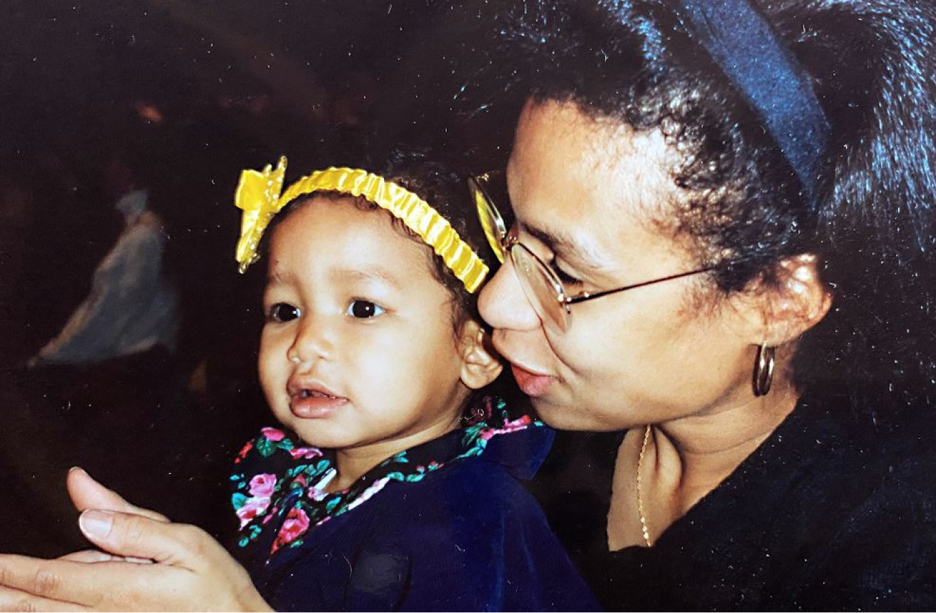 A nostalgic photo of Edith Cooper and a young Jordan Taylor. Edith is wearing glasses and a dark outfit, leaning close to Jordan, who is a toddler wearing a floral dress and a yellow headband. The warm moment captures their bond, set against a softly blurred background.