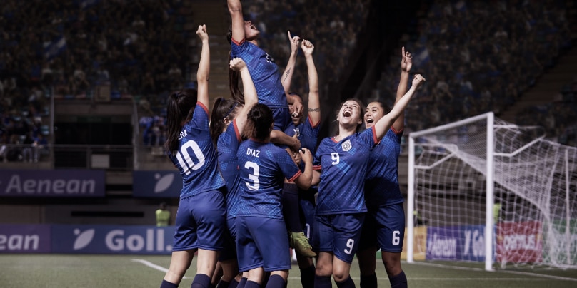 A group of female soccer players in blue and red uniforms celebrating a victory. They lift a teammate into the air, who shouts with joy, surrounded by teammates cheering and raising their arms in triumph.