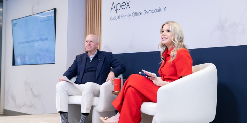 A man dressed in blue, and a woman dressed in red sit on stage at an event, looking out toward the audience. A sign behind them says, “Apex Global Family Office Symposium”.  