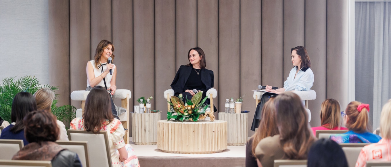 A panel discussion featuring three women, from left to right, Candace Nelson, Jessica Farron, and Ling Pong, on stage in a modern and elegant setting. Candace Nelson, wearing a white sleeveless top and patterned skirt, speaks into a microphone while smiling. Jessica Farron, in a black outfit, listens attentively. Ling Pong, dressed in a light blue blouse, holds a microphone and notes as she engages in the conversation. The stage is decorated with soft beige paneling, wooden tables, and floral arrangements while an audience of women observes.