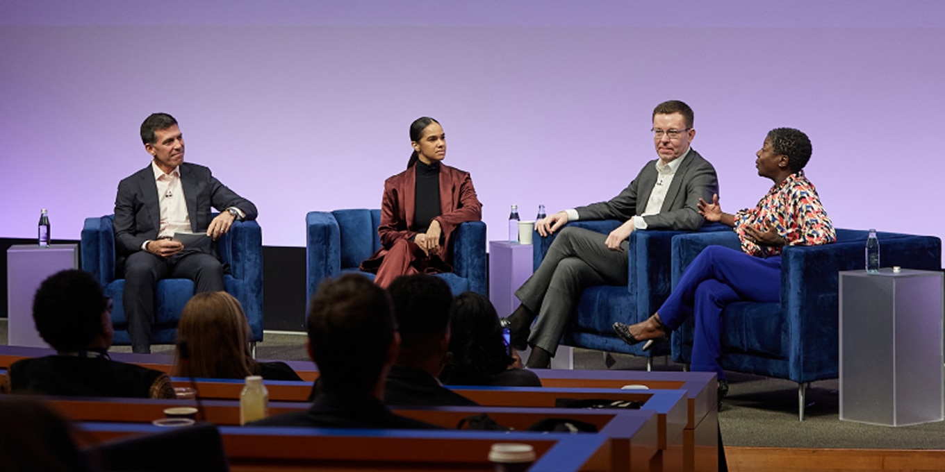 John Waldron, Misty Copeland, Henry Timms, and Thelma Golden participate in a panel discussion at the Rising Leaders Forum. They are seated on a stage in deep blue chairs, engaged in conversation. Thelma Golden gestures expressively while speaking, as the other panelists listen attentively. The audience watches from tiered seating in the foreground.