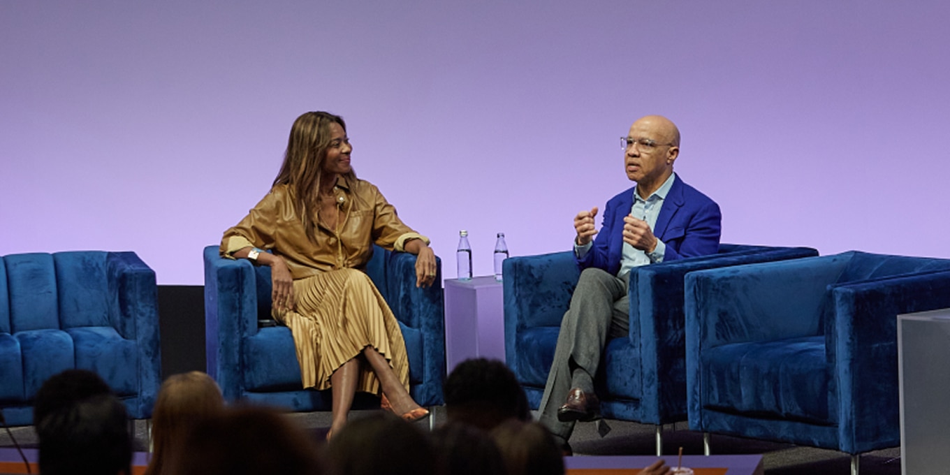 Asahi Pompey and Darren Walker engage in a discussion on stage at the Rising Leaders Forum. Asahi Pompey, wearing a gold-toned pleated dress, listens attentively while Darren Walker, dressed in a blue blazer, gestures expressively as he speaks. They are seated in deep blue chairs against a purple-lit background, with an audience watching from the foreground.
