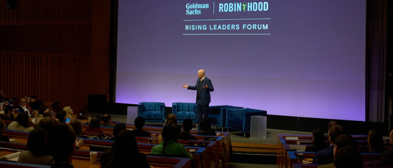 Goldman Sachs Chairman and CEO David Solomon speaks on stage at the Rising Leaders Forum, hosted in collaboration with Robin Hood. He gestures with open arms as he addresses an audience seated in a modern auditorium with a large screen displaying the event branding behind him.