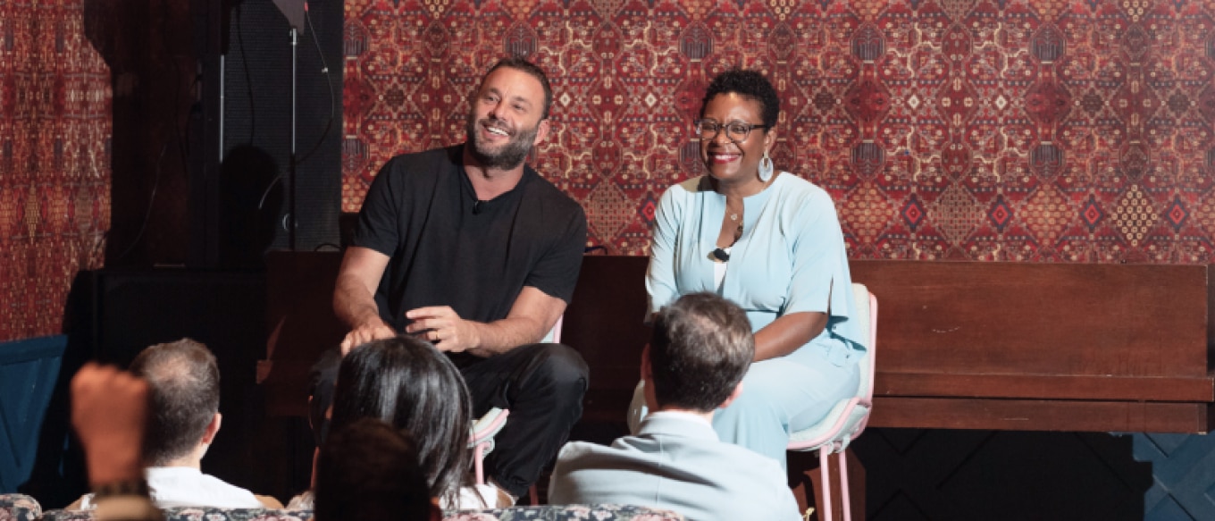 David Grutman and Nicole Pullen Ross seated on a stage engaging with an audience. David Grutman, on the left, is dressed in a black outfit, smiling warmly, while Nicole Pullen Ross, on the right, wears a light blue outfit and also smiles. The background features a patterned decorative wall, and the setting has a relaxed and conversational atmosphere.