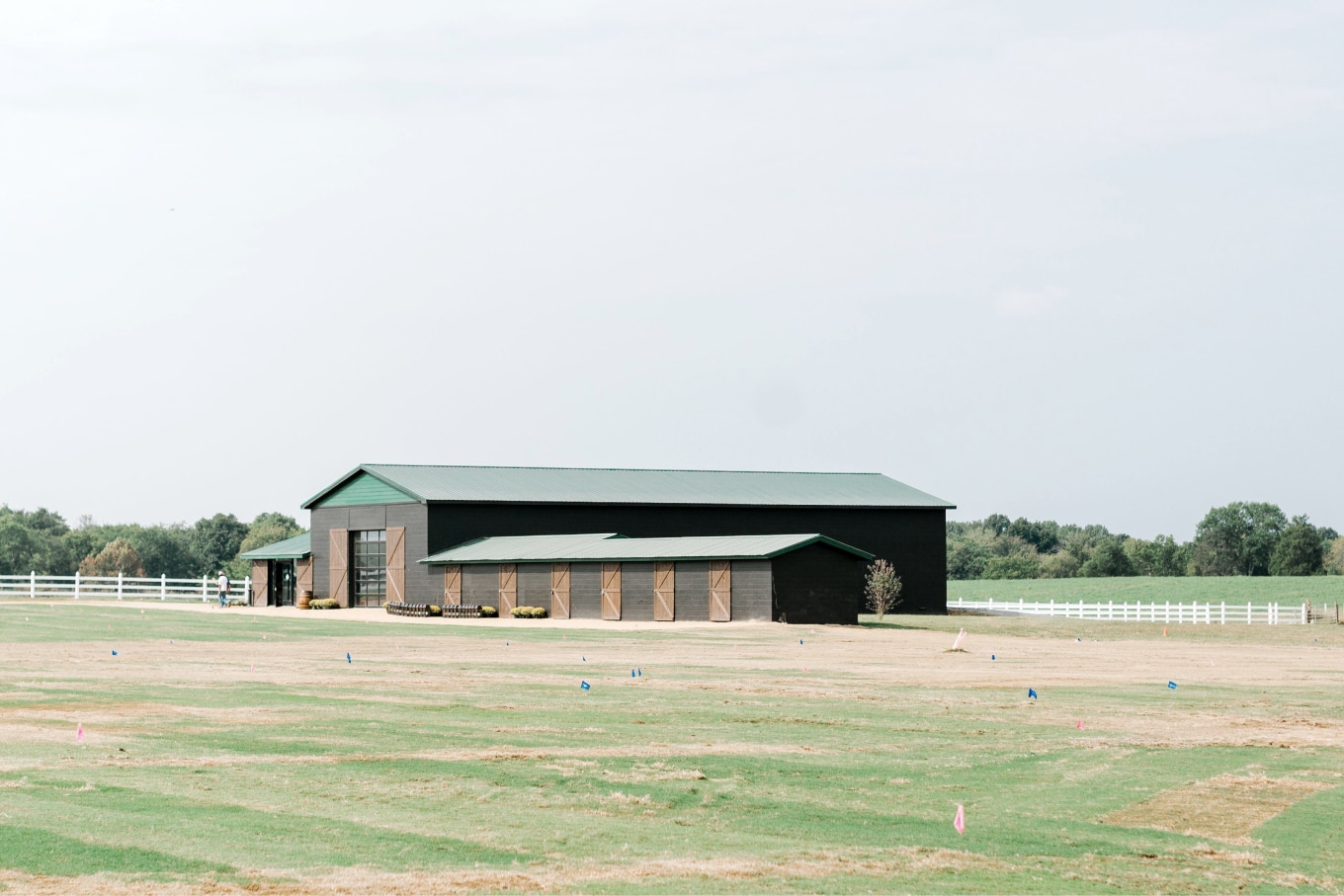 A wide view of the Nearest Green Distillery property showing a large black barn with a green roof, surrounded by a vast open field and a white fence. The scene conveys a serene and spacious environment under a clear sky.