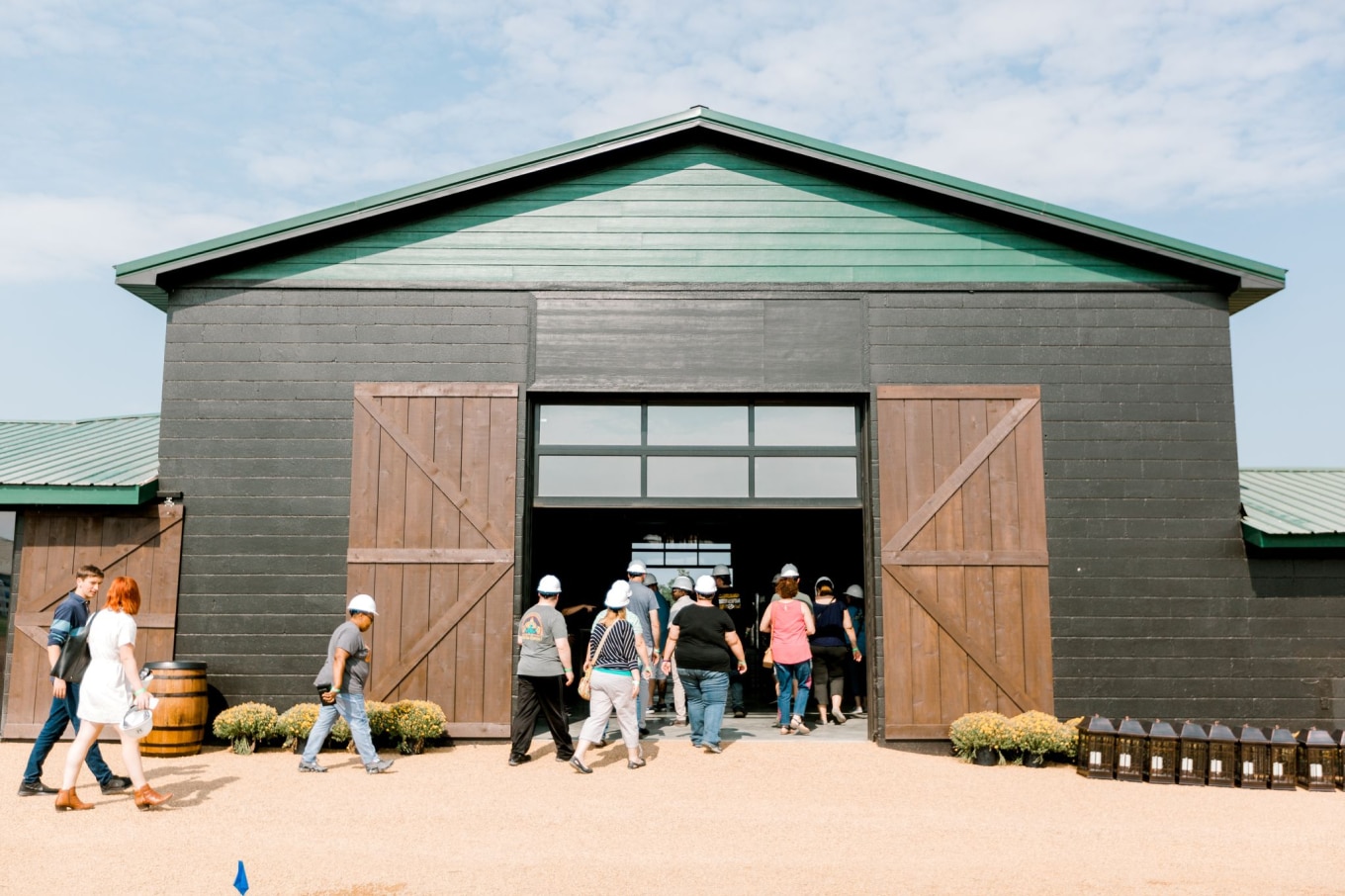 A group of people wearing hard hats walking into a large black barn with wooden doors wide open. The barn has a green roof, and the surrounding area features gravel, small plants, and barrels, set under a bright blue sky.