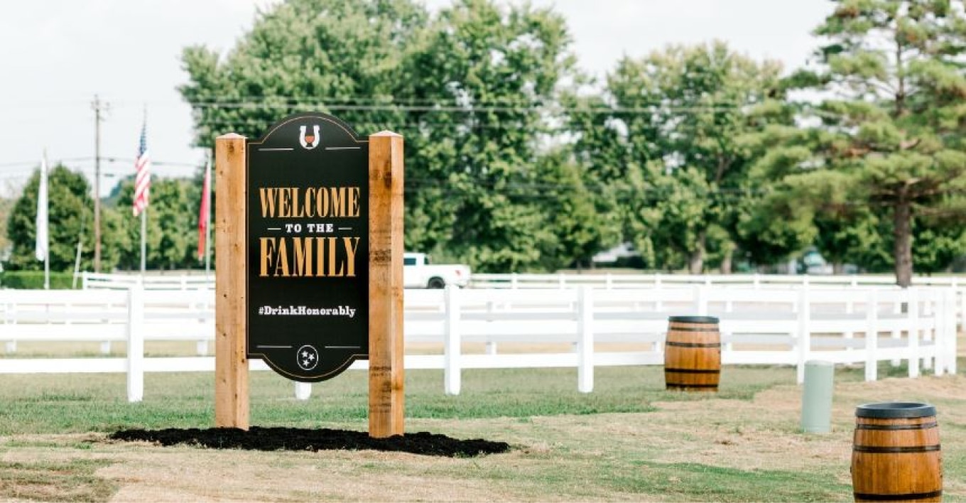 A welcoming sign reading 'Welcome to the Family' with the hashtag #DrinkHonorably at the Nearest Green Distillery. The area is surrounded by white fences, wooden barrels, and trees, creating an inviting and scenic atmosphere.