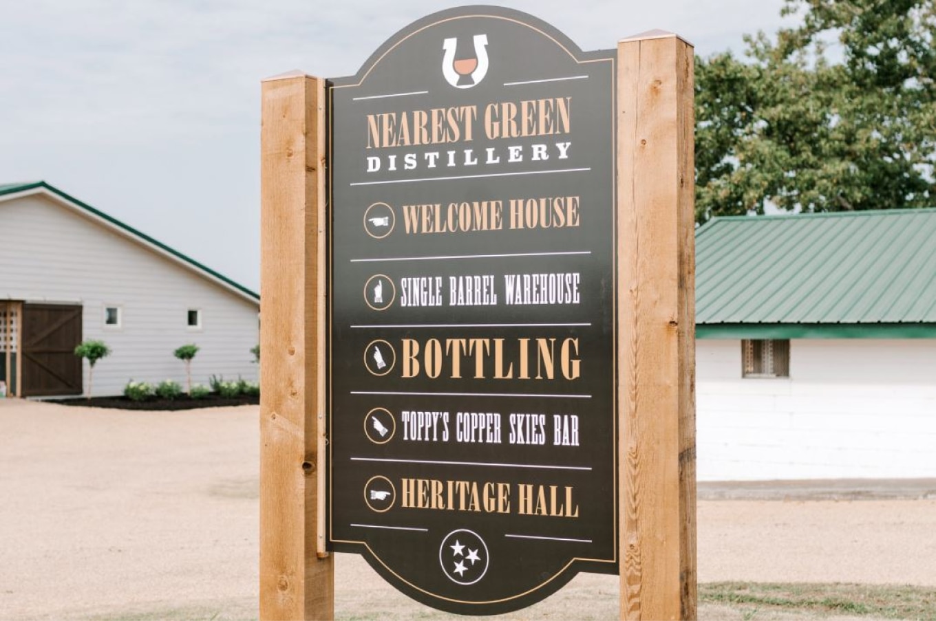 A wooden sign for the Nearest Green Distillery featuring directions to various locations on the property, including the Welcome House, Single Barrel Warehouse, Bottling, Toppy's Copper Skies Bar, and Heritage Hall. The background includes a white building with a green roof and surrounding plants.
