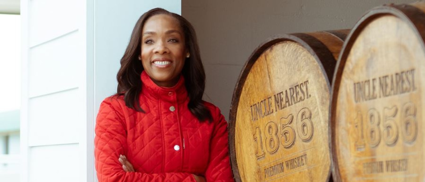 Fawn Weaver standing confidently next to large wooden barrels labeled 'Uncle Nearest 1856 Premium Whiskey.' She is wearing a red quilted jacket and smiling warmly, with a bright and inviting background that suggests a distillery or outdoor setting.