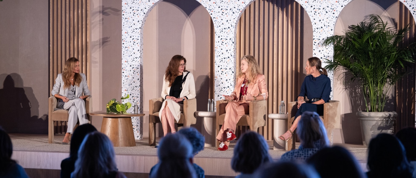A panel discussion featuring four women on a stage with a modern, elegant backdrop. The panelists are engaged in conversation, with one speaking while the others listen attentively. The audience is visible in the foreground.
