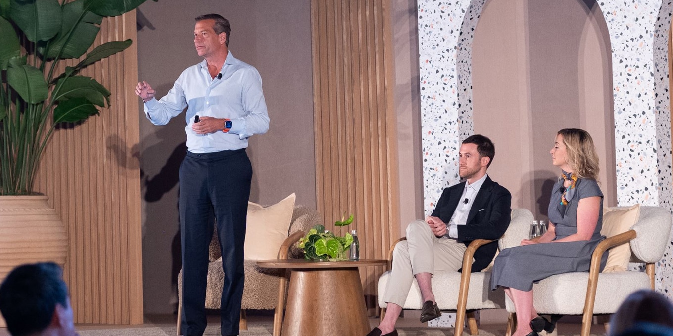 A man in a light blue dress shirt and dark pants stands on stage, delivering a presentation while holding a microphone. Two panelists sit attentively on modern wooden chairs: a man in a dark blazer and light-colored pants and a woman in a gray dress with a patterned scarf. A small wooden table with greenery and bottled water sits between them. The stage features warm wood paneling, terrazzo archways, and lush plants
