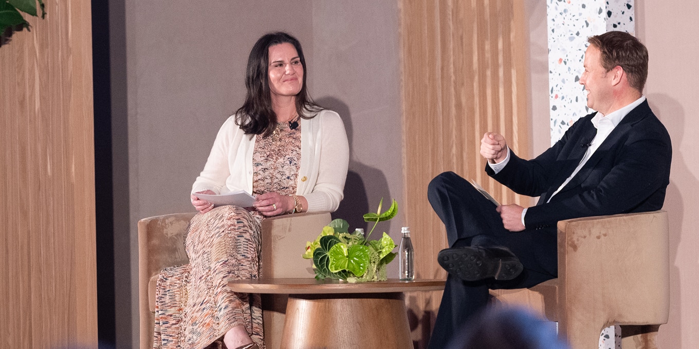 A woman and a man are seated on a stage engaged in a discussion. The woman, wearing a patterned dress with a white cardigan, holds papers and smiles while listening. The man, dressed in a dark suit and white shirt, gestures as he speaks. They are seated in modern beige chairs with a small wooden table between them, decorated with greenery and bottled water. The stage has warm wooden paneling and terrazzo accents.