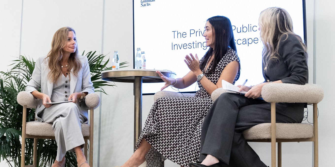 Three women are seated in a panel discussion in a professional setting. The woman on the left, dressed in a light gray suit, listens attentively while holding a notepad. The woman in the center, wearing a patterned dress, gestures as she speaks. The woman on the right, dressed in a dark suit, takes notes while conversing. A round table with bottled water is placed between them, and a screen behind them displays the topic: "The Private & Public Investing Landscape." The setting is modern, with neutral tones and greenery.