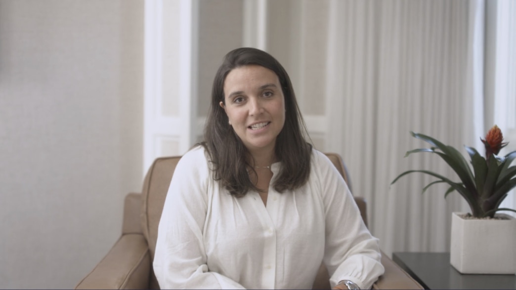 Saara Pritchard sits wearing a white blouse sits in a brown armchair, speaking to the camera with a friendly expression. The background features a neutral-toned room with white panel doors, curtains, and a potted plant with red flowers on a side table.