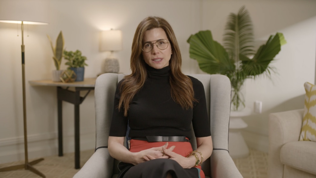 Desiree Gruber seated in a modern, well-lit room with warm decor. She is wearing a black turtleneck, large glasses, and a red skirt with a black belt, accessorized with gold bracelets. The background includes a lamp, plants, and a calming, stylish atmosph