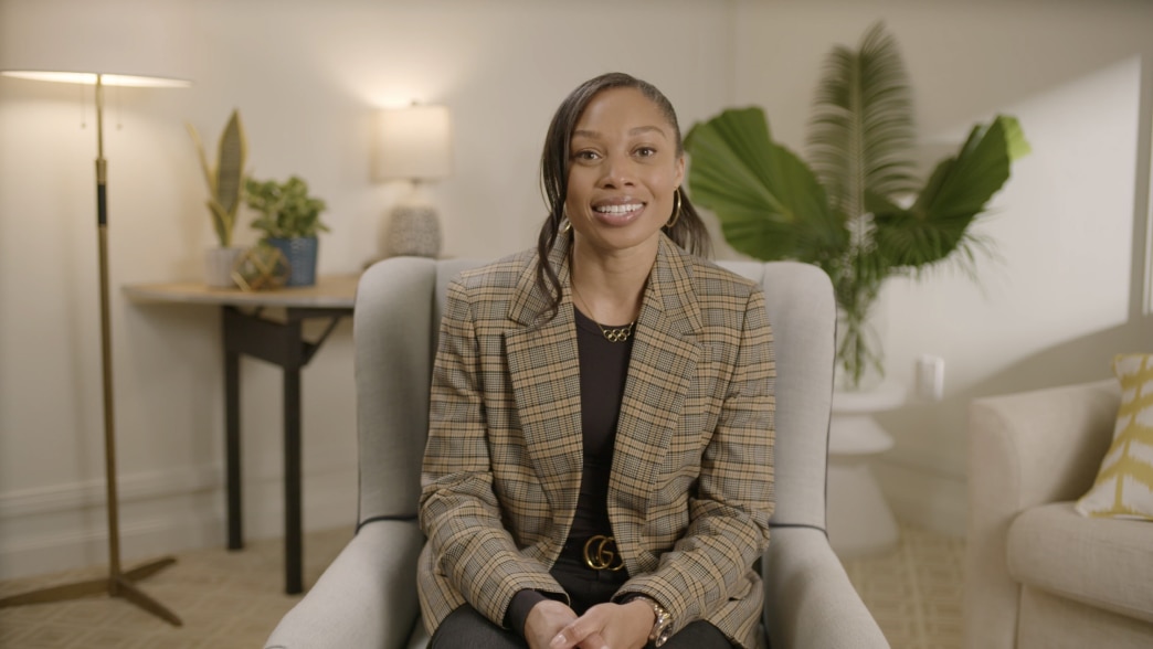 Allyson Felix seated in a modern, warmly lit room with neutral decor. She is wearing a stylish plaid blazer over a black top, accessorized with gold jewelry, and is smiling confidently at the camera. The background features a lamp, plants, and a calm, inv