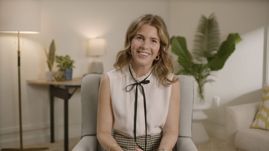 Candace Nelson sitting in a well-lit, modern room with a warm and inviting atmosphere. She is wearing a sleeveless white blouse with a black ribbon tie and a patterned skirt, smiling warmly at the camera. The background features a lamp, a table with plant