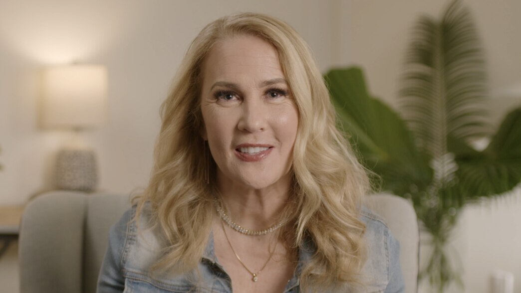 An image from a video featuring Jennifer Aaker speaking in a well-lit, cozy setting. She has a warm and engaging expression. The background includes a softly lit lamp and green plants.