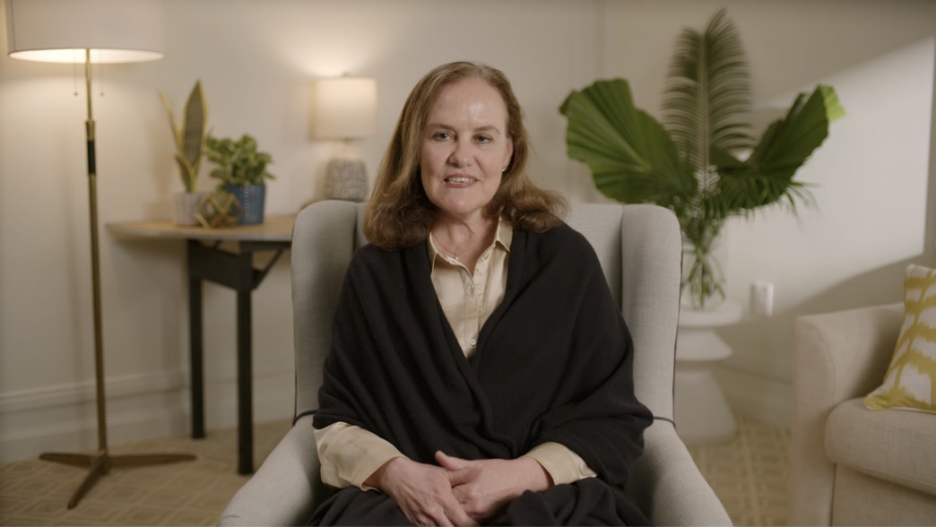 Michele Flournoy sitting in a comfortable armchair, speaking in a video. She is wearing a beige blouse with a black shawl draped over her shoulders. The background features a warmly lit room with plants and a modern interior design.