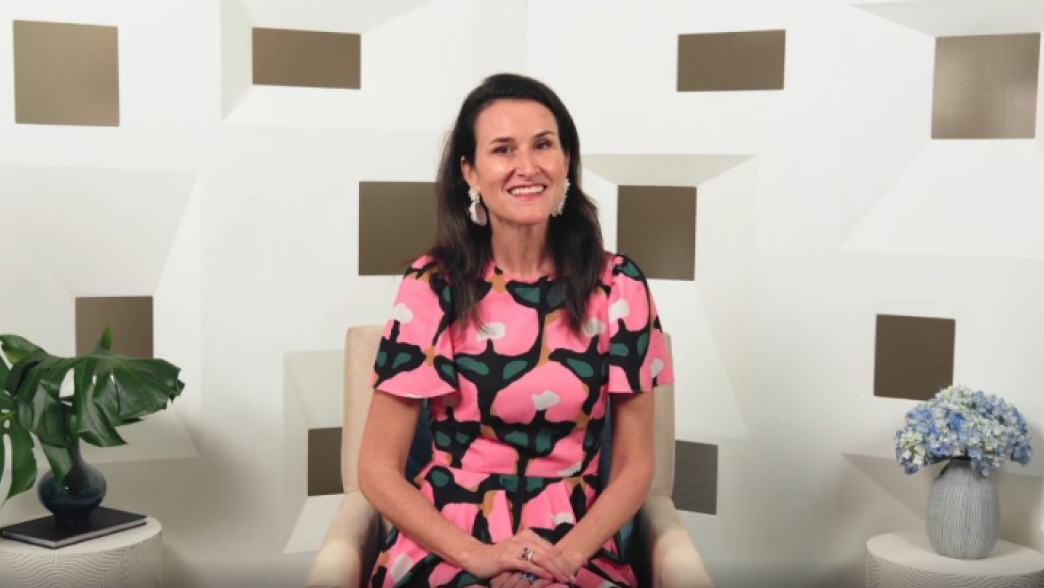 Lydia Fenet speaking in a video, seated in an armchair against a geometric patterned backdrop. She is wearing a vibrant pink and black patterned dress and smiling. The backdrop features a modern geometric design with muted gold and white tones, along with