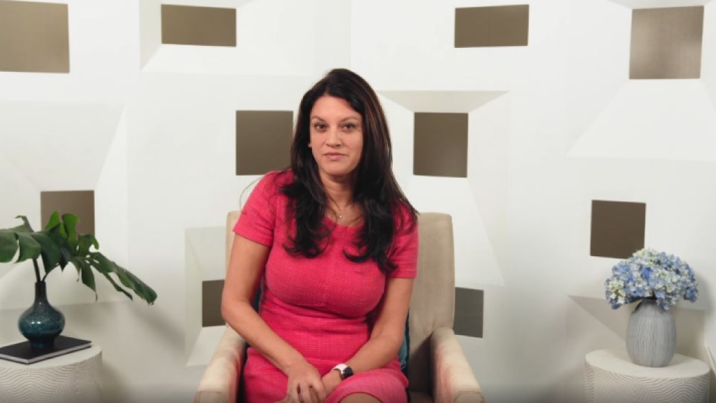 Navyn Salem seated in a chair, looking directly at the camera during a video presentation. She is wearing a bright pink dress with a confident expression. The backdrop features a modern geometric design with white and muted gold tones, complemented by dec
