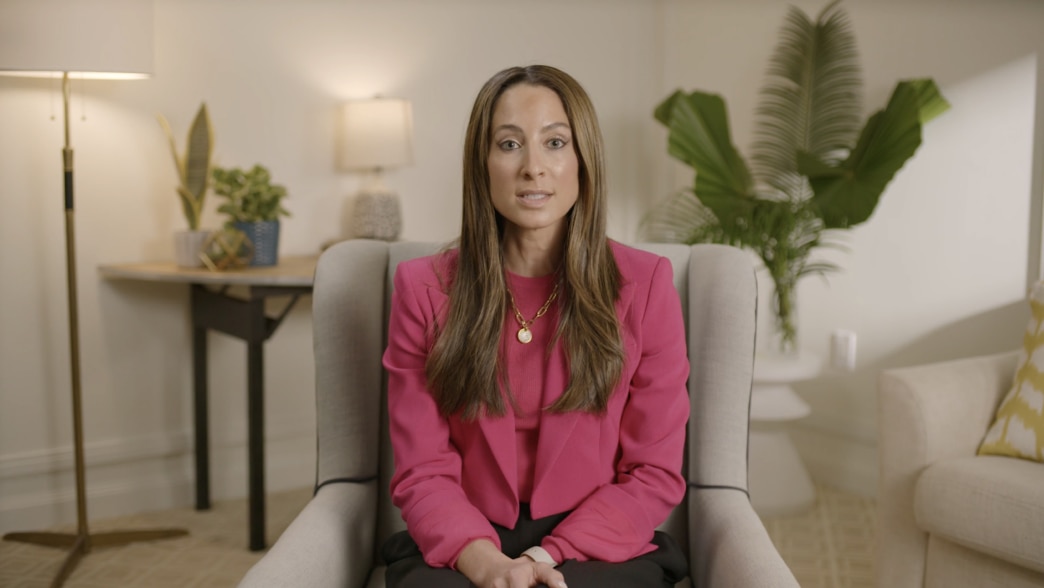 An image from a video featuring Alexandra Idol seated in a modern, well-lit setting. She is wearing a vibrant pink blazer over a matching top, accessorized with a gold chain necklace. She has a poised and professional expression while speaking. The backgr