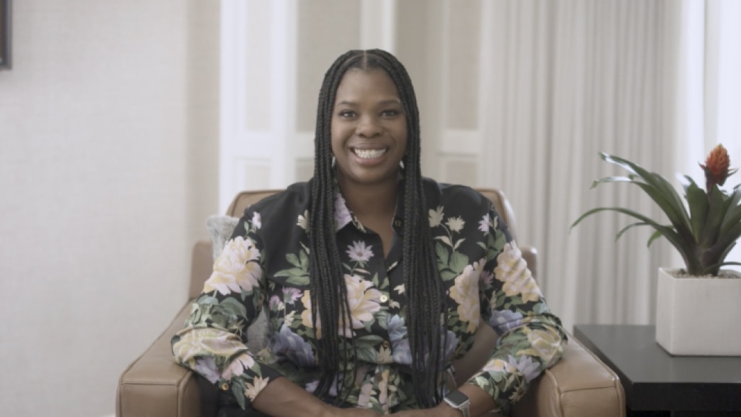 Jasmine Crowe-Houston smiling warmly while seated in a bright room with neutral decor. She is wearing a floral-patterned blouse and has long braided hair, creating a friendly and approachable atmosphere.