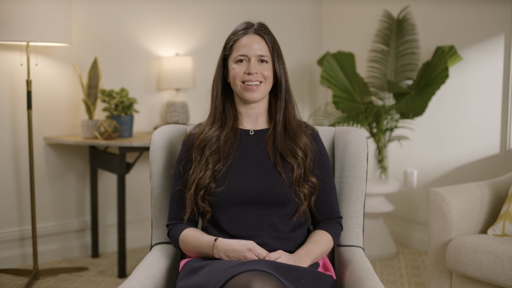 Rachel Knaster seated in a light gray armchair, looking directly at the camera with a smile during a video presentation. She is wearing a navy blue dress with a pink accent and has long, wavy brown hair. The background features a well-lit, modern interior