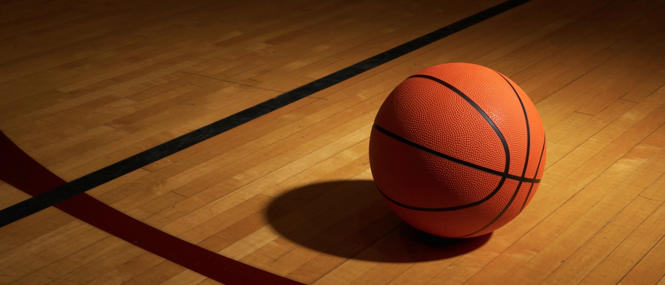 A basketball resting on a polished wooden court, casting a shadow under dramatic lighting. The court's boundary lines are visible in the background.