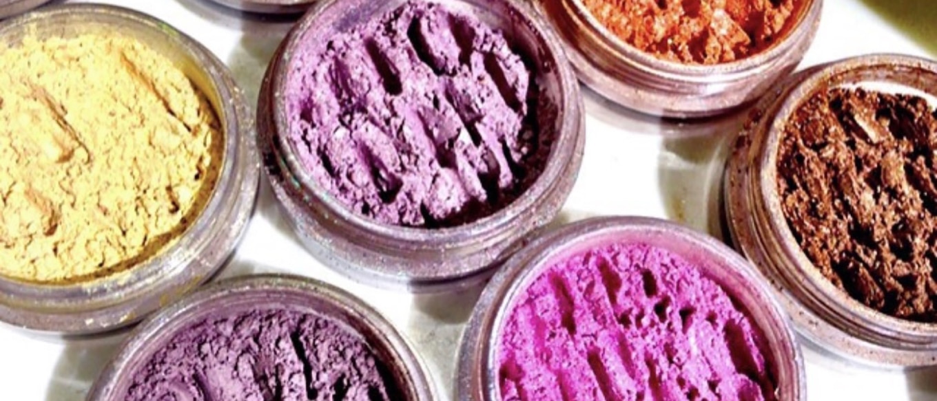 A collection of colorful loose powder pigments displayed in small round containers. The colors include yellow, various shades of purple, pink, orange, and brown, showcasing a textured, finely milled appearance.