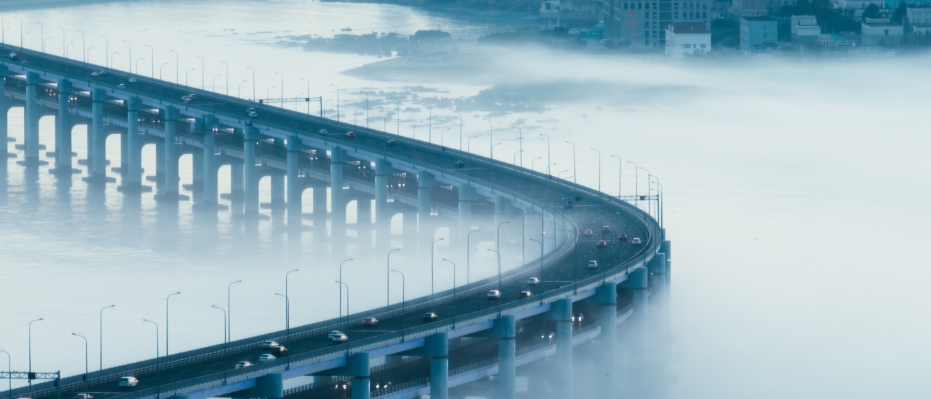 A bridge curving over a body of water, partially shrouded in mist. The bridge has multiple lanes with cars moving in both directions, and it is supported by large pillars extending into the water. The fog creates a dreamy atmosphere, blending the water and sky, while a cityscape with buildings is faintly visible in the background