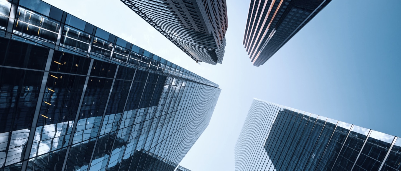 A low-angle photograph of modern skyscrapers towering towards the sky. The buildings have reflective glass facades and sleek architectural designs, creating a sense of depth and scale. The sky is a clear blue, contrasting with the structures' geometric lines and mirrored surfaces. The perspective emphasizes the dominance of urban architecture in a financial or business district.