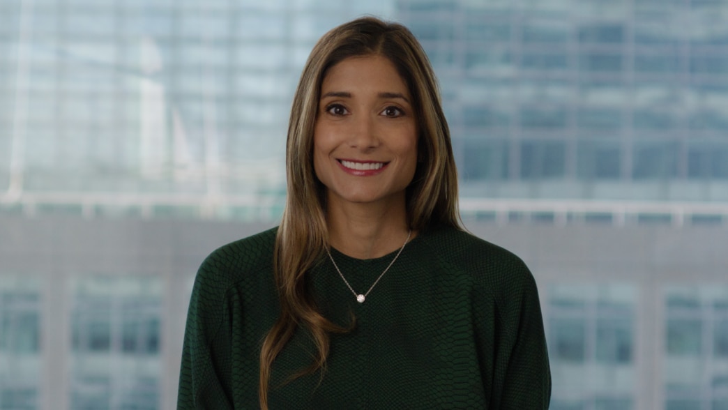 A professional portrait of Meena Flynn, smiling at the camera. She is wearing a dark green textured blouse and a silver necklace with a small pendant. Her long, light brown hair is styled naturally. The background features a blurred modern glass office bu