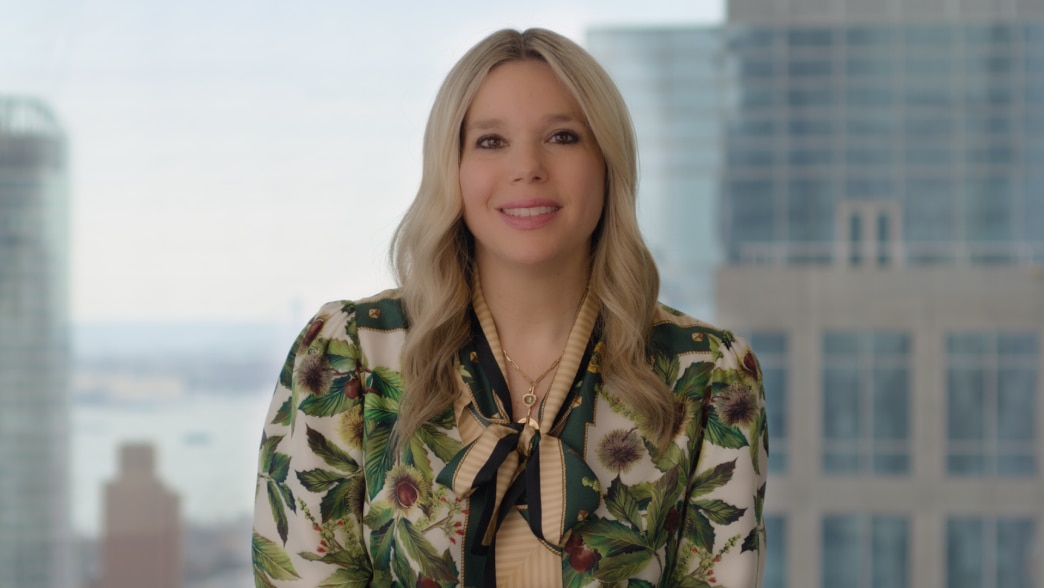 Video static - A professional portrait of Naison Tarajano, smiling at the camera. She has long, wavy blonde hair and is wearing a stylish blouse with a green and gold botanical print, featuring a bow detail at the neckline. She also wears a gold pendant n
