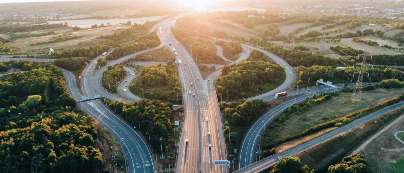 Aerial view of a complex highway interchange surrounded by lush greenery, with vehicles moving along the roads under a golden sunset.