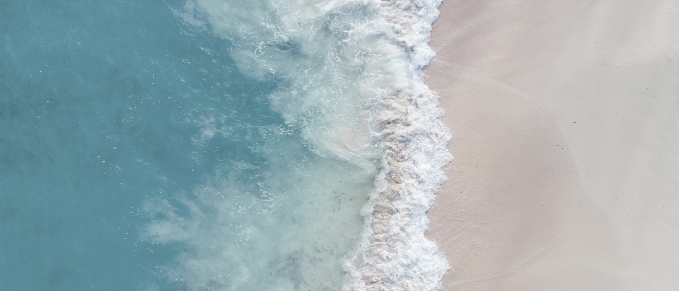 An aerial view of ocean waves gently washing onto a soft, sandy beach. The deep blue water transitions into frothy white foam as it meets the shoreline, contrasting the sea and sand. The natural textures and colors capture the rhythmic movement of the waves, evoking a sense of serenity and relaxation.