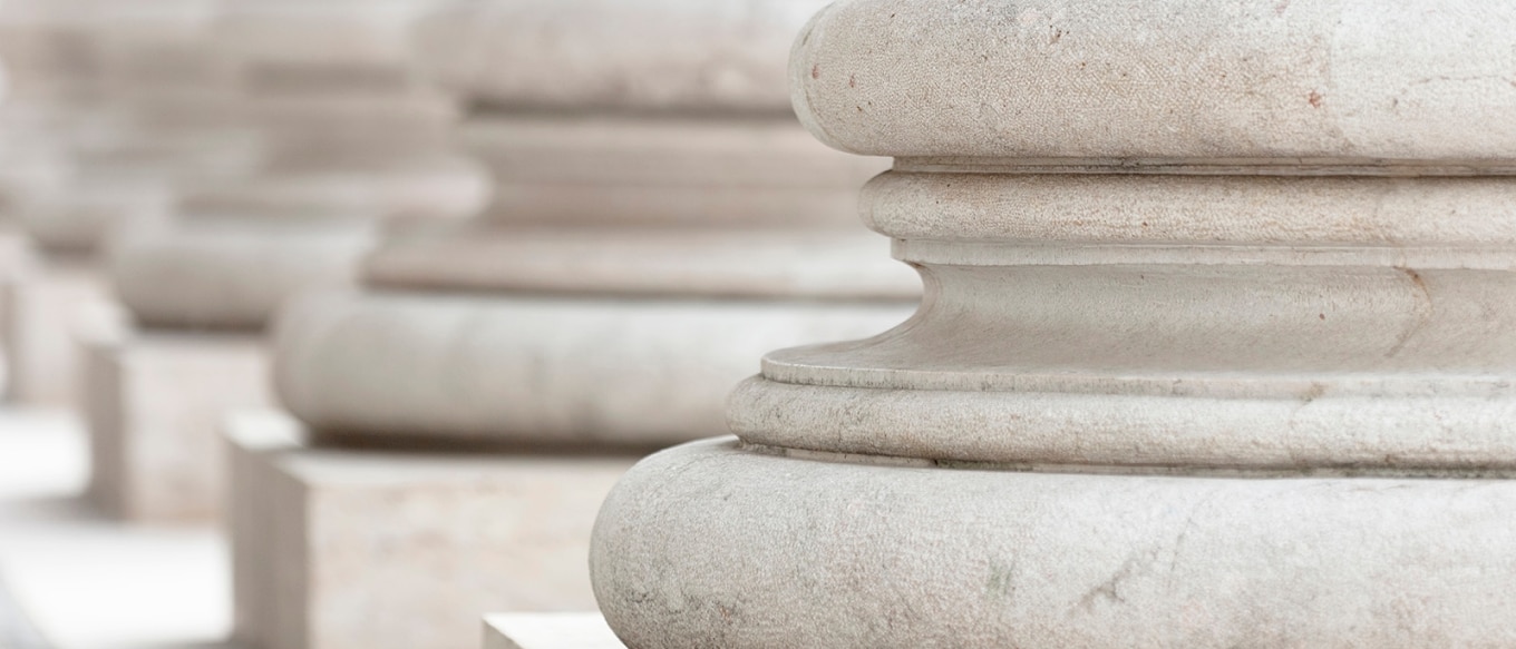 A close-up photograph of the sturdy stone bases of classical architectural columns. The smooth, rounded design of the columns showcases intricate craftsmanship, symbolizing strength, stability, and tradition. The soft lighting and neutral tones highlight the texture of the stone, while the repetition of the columns creates a sense of depth and perspective.