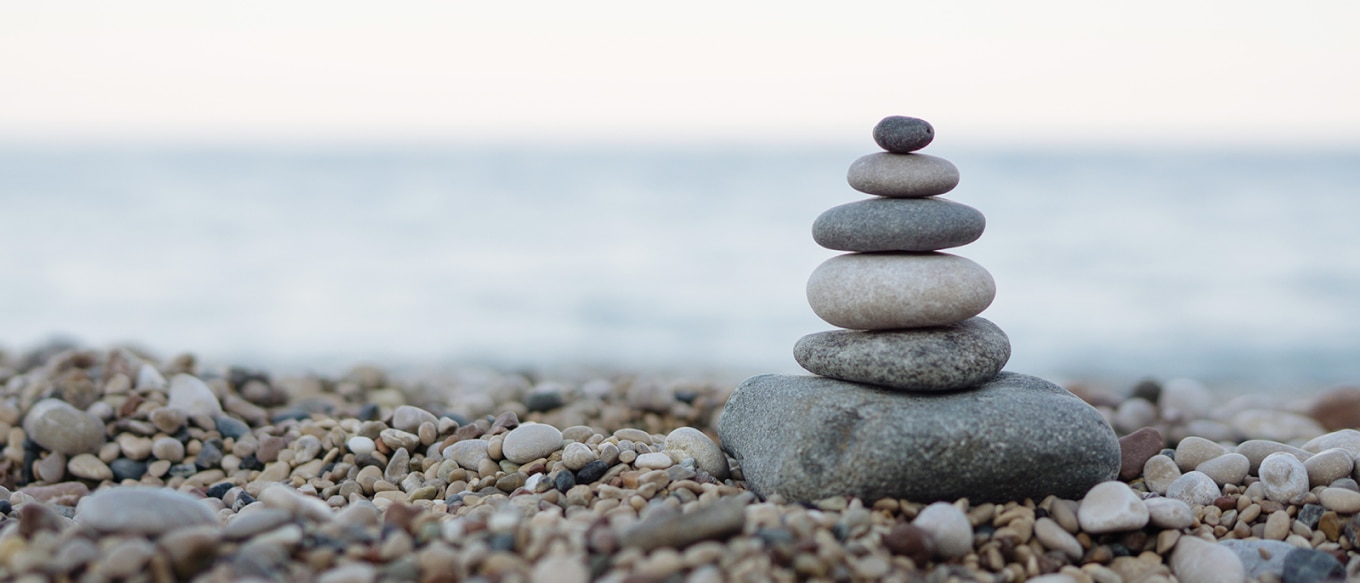 A beach scene featuring a small, carefully balanced stack of smooth pebbles on a rocky shoreline. The background showcases a calm, blurred ocean meeting the sky at the horizon, creating a minimalist composition. The soft, natural lighting enhances the neutral tones of the stones, evoking a sense of tranquility, balance, and mindfulness.