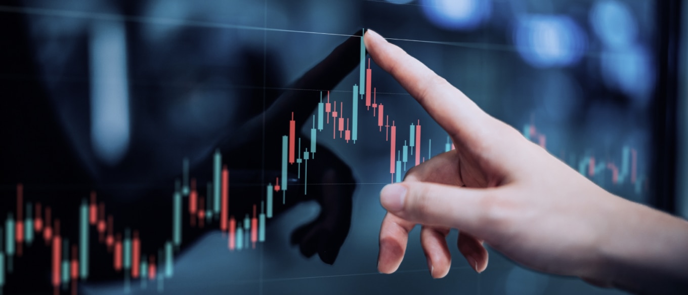 A close-up of a hand pointing at a digital financial chart displayed on a screen. The chart features red and green candlestick bars, indicating market trends and fluctuations. The hand's reflection is visible on the glossy surface, adding depth to the image. The background is blurred with cool-toned lighting, creating a modern and high-tech financial setting.