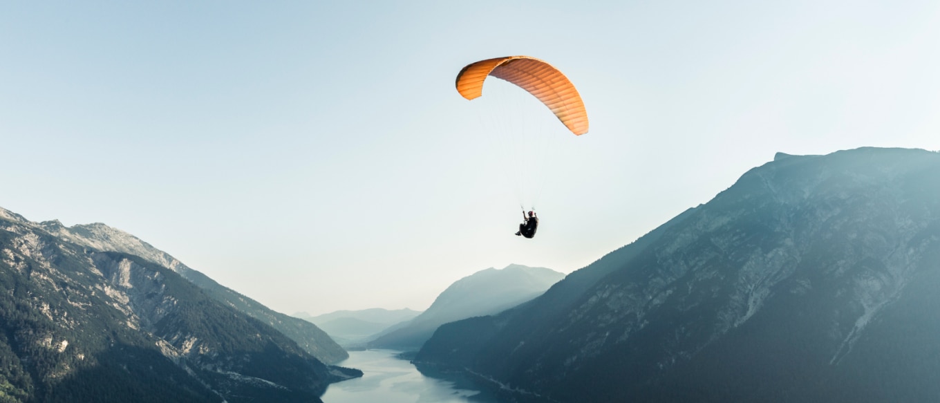 A photograph of a paraglider soaring high above a serene mountain landscape. The orange canopy contrasts against the soft blue sky, while the rugged mountains and a reflective lake stretch into the distance below.
