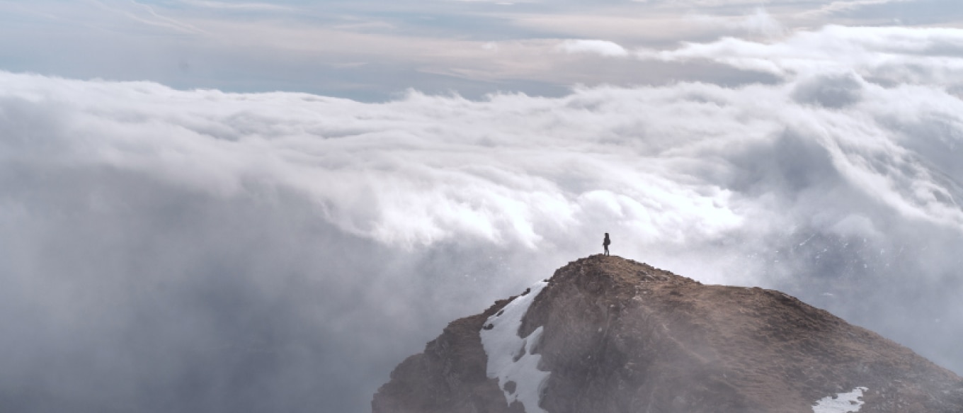 A landscape featuring a lone figure standing atop a rugged mountain peak, surrounded by clouds. The vast expanse of clouds below creates a sense of height and isolation while patches of snow cling to the rocky terrain. The sky above mixes soft blues and grays, adding to the atmosphere.