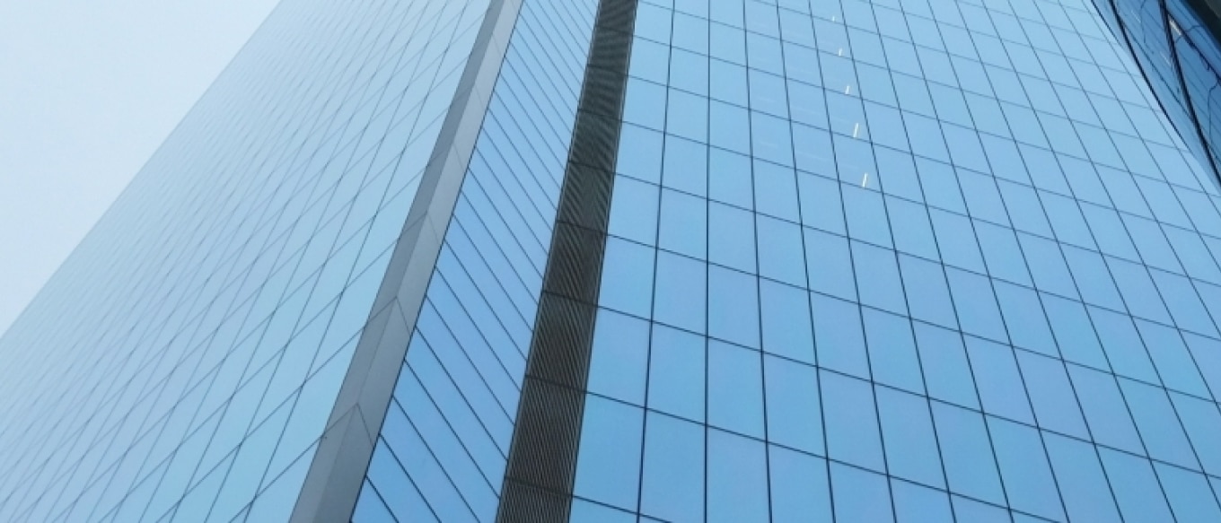 A low-angle view of a modern glass skyscraper reflecting the sky. The building's reflective blue-tinted windows create a geometric pattern