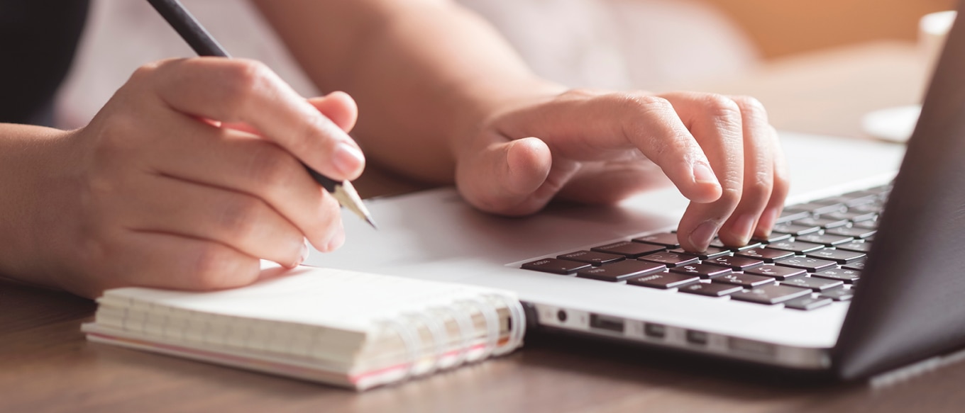 A person working at a desk using a laptop, with one hand typing on the keyboard and the other holding a pen over an open notebook. The scene is lit by warm natural light, creating a focused and productive atmosphere.