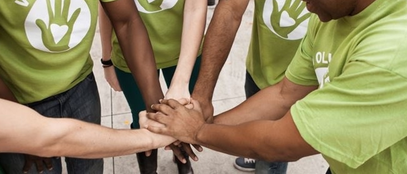 A diverse group of volunteers wearing matching green t-shirts with a hand and heart logo join hands in a team huddle.